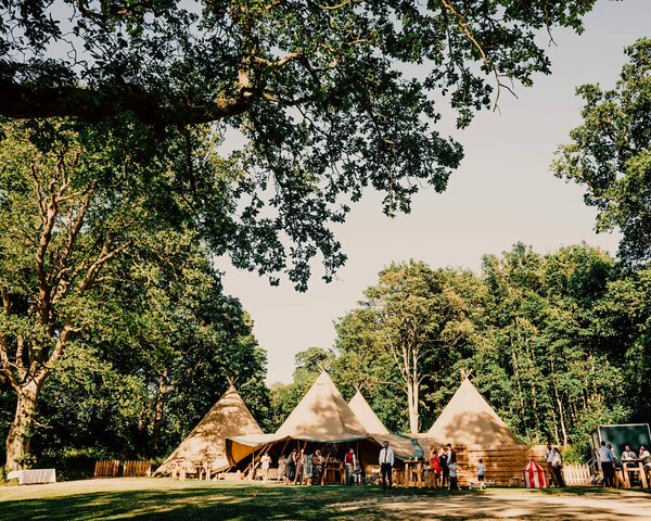 Four tipis sit amongst ancient woodland with a magical lake in the forefront, wedding guests are standing outside celebrating